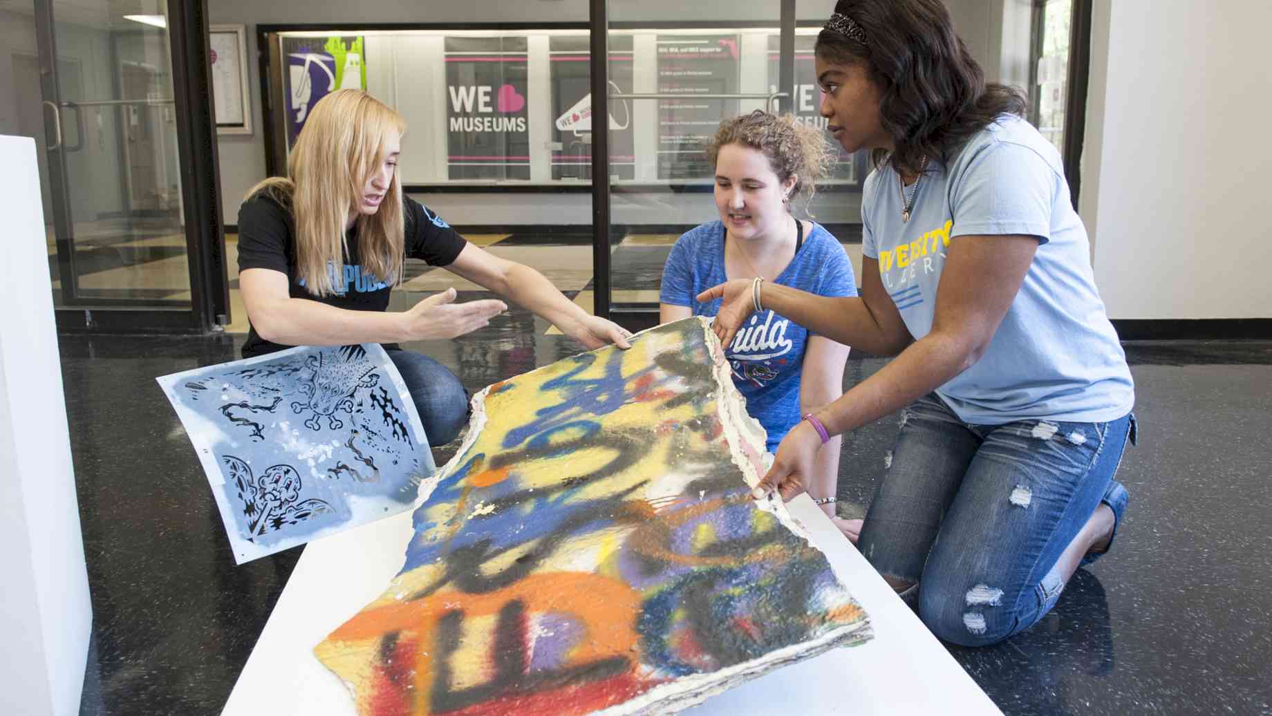 Museums Studies students install art in a gallery. [Alt text: Three students kneel in an art gallery, holding pieces of art and speaking.]